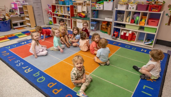 A group of young children sit on a colorful alphabet rug in their classroom, surrounded by shelves of toys. Many of the children are smiling at the camera.