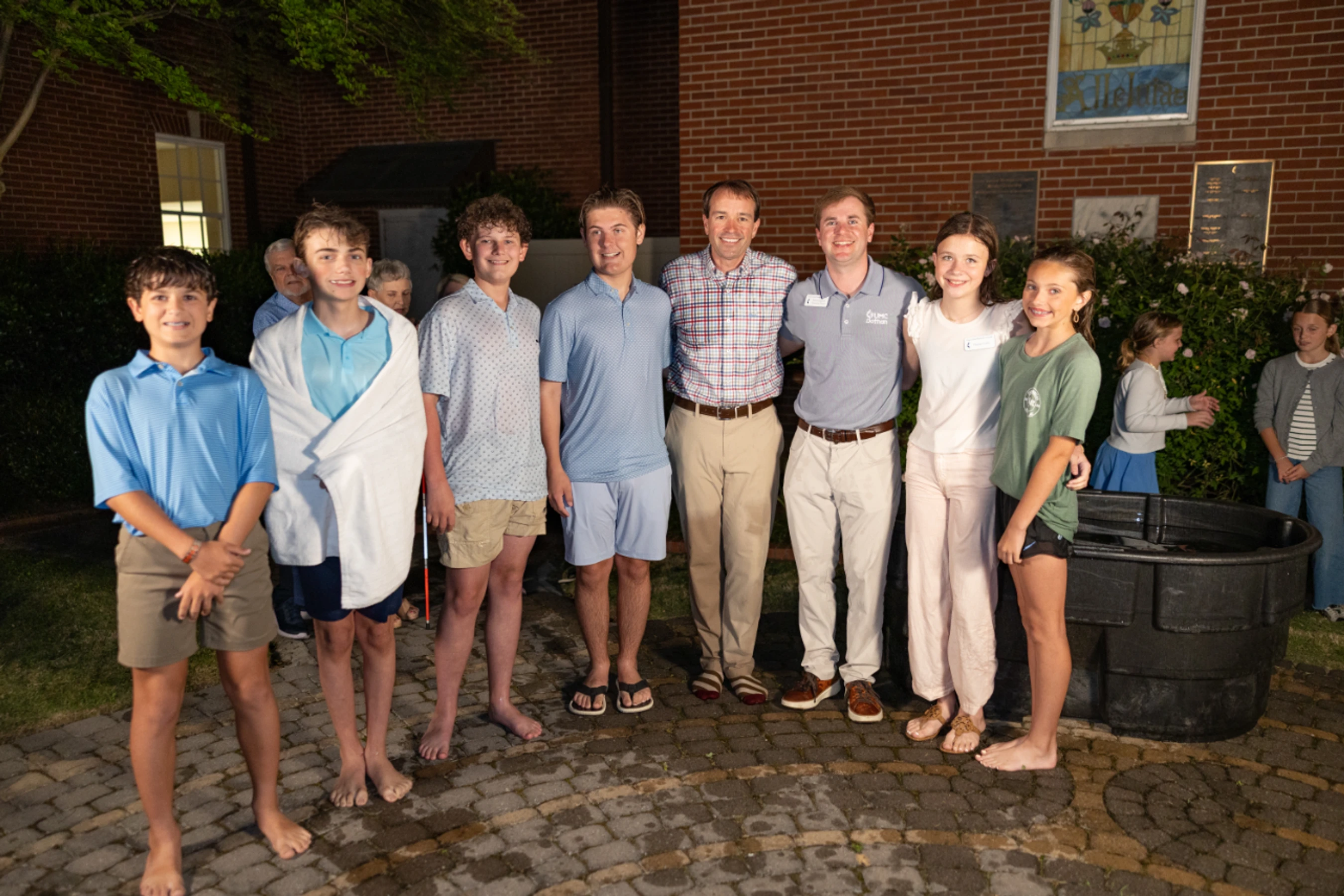 A group of smiling teens and adults pose for a photo on a brick patio at night, with one boy in a towel standing near a large tub of water.