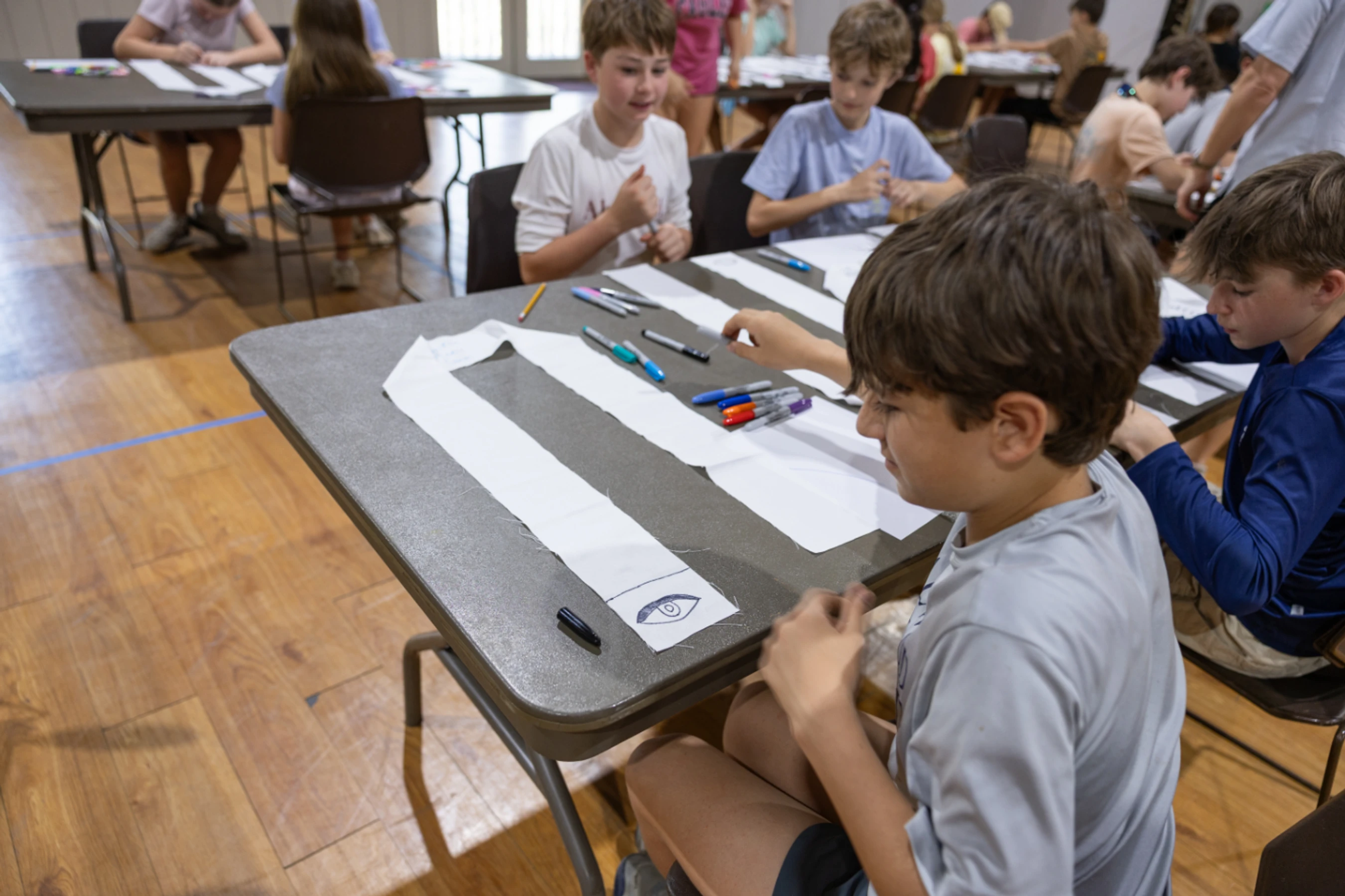 A group of young children sit at tables in a large room, using colored markers to draw designs on long, white fabric stoles as part of a creative activity.