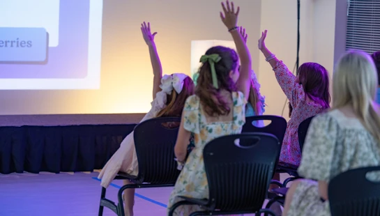 A view from behind a group of young girls sitting on chairs, eagerly raising their hands while watching a presentation on a lit stage.