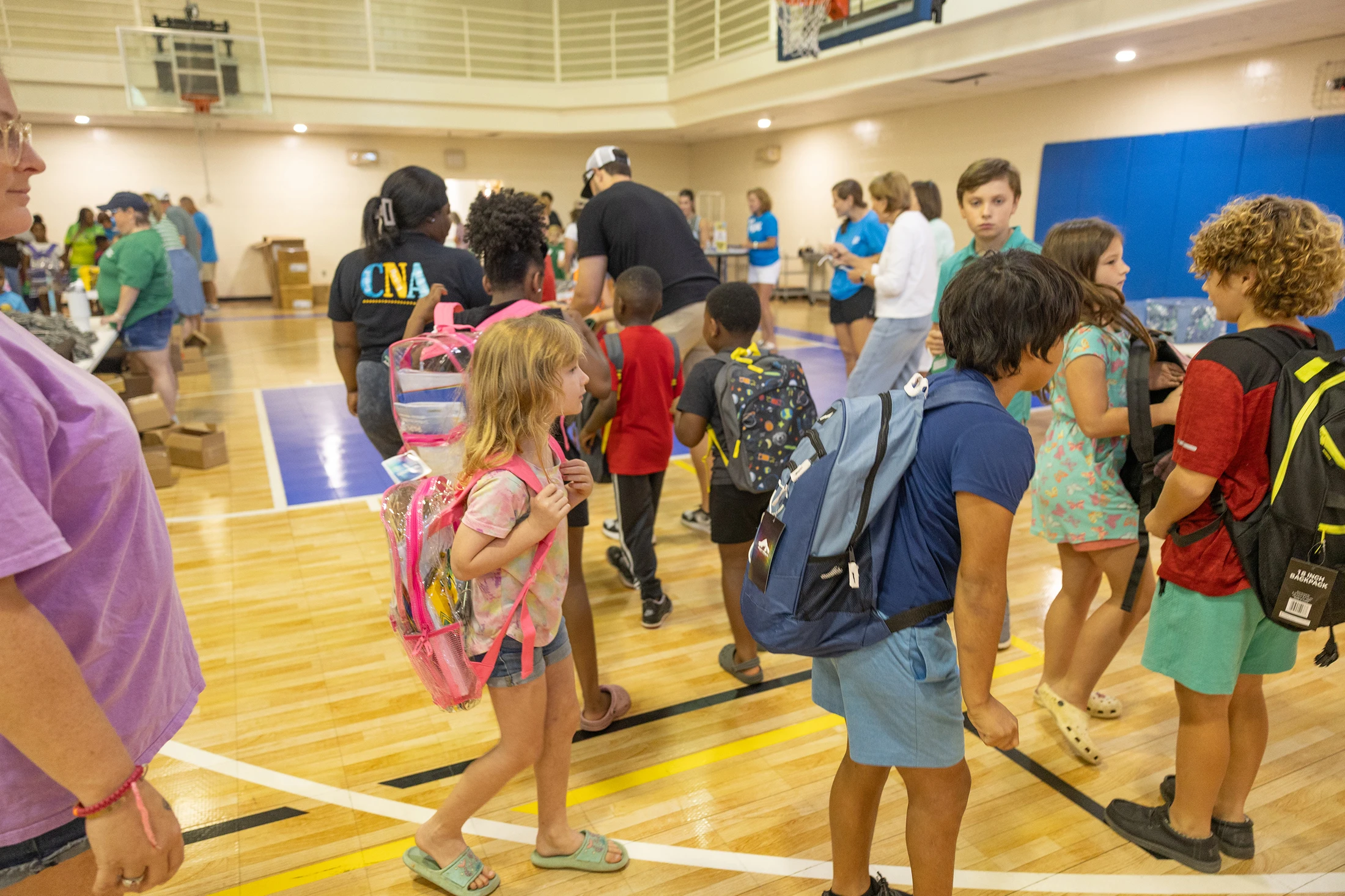A diverse group of children wearing new backpacks stand in line inside a school gymnasium during a community back-to-school supply drive.