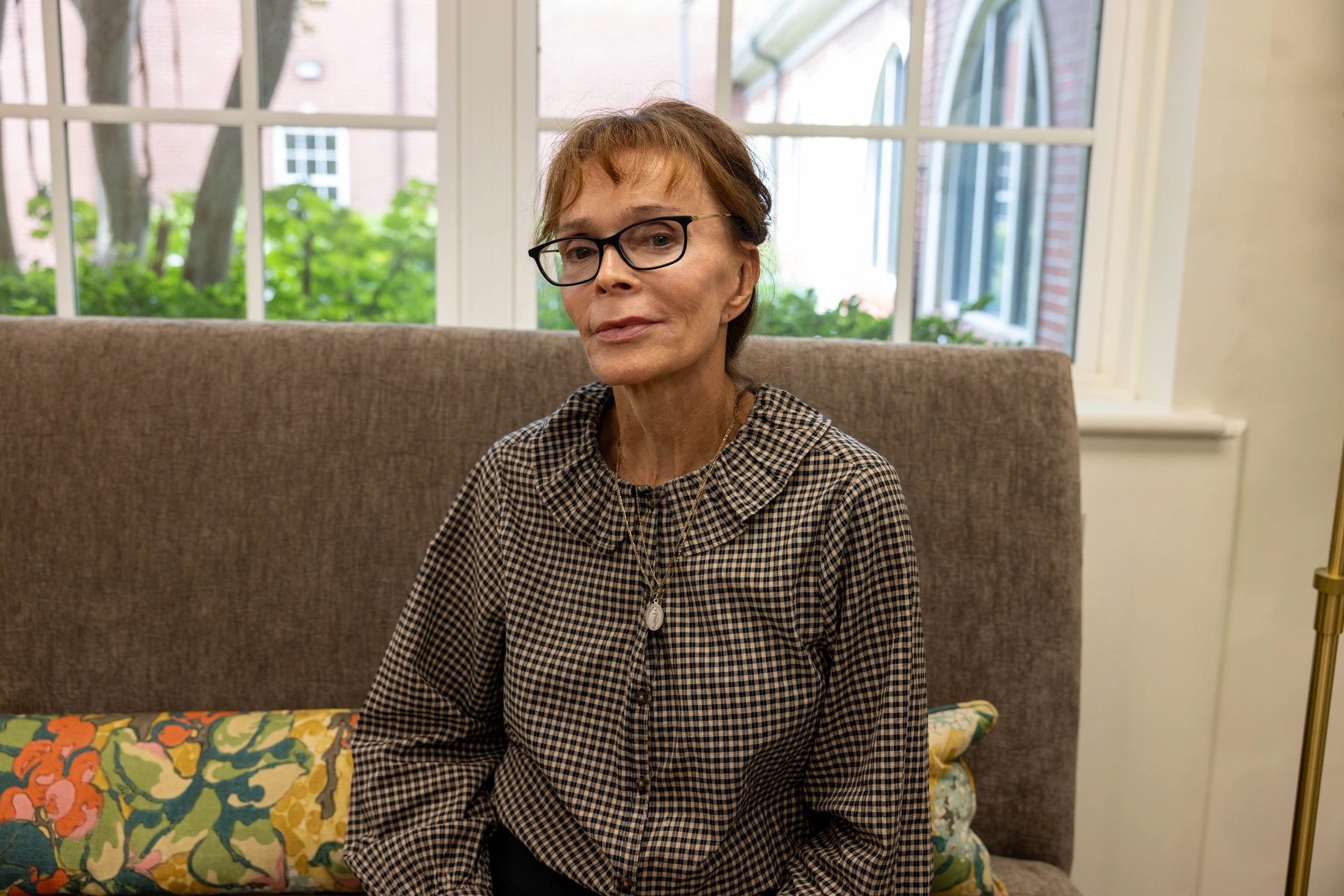 A woman with reddish-brown hair and glasses, wearing a checkered shirt, sits on a brown couch in front of a large window and looks at the camera.