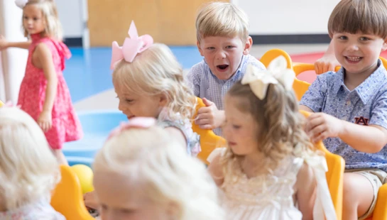 A group of young children, mostly blonde, sit in a yellow toy train indoors. One boy shouts with excitement while another boy smiles at the camera.