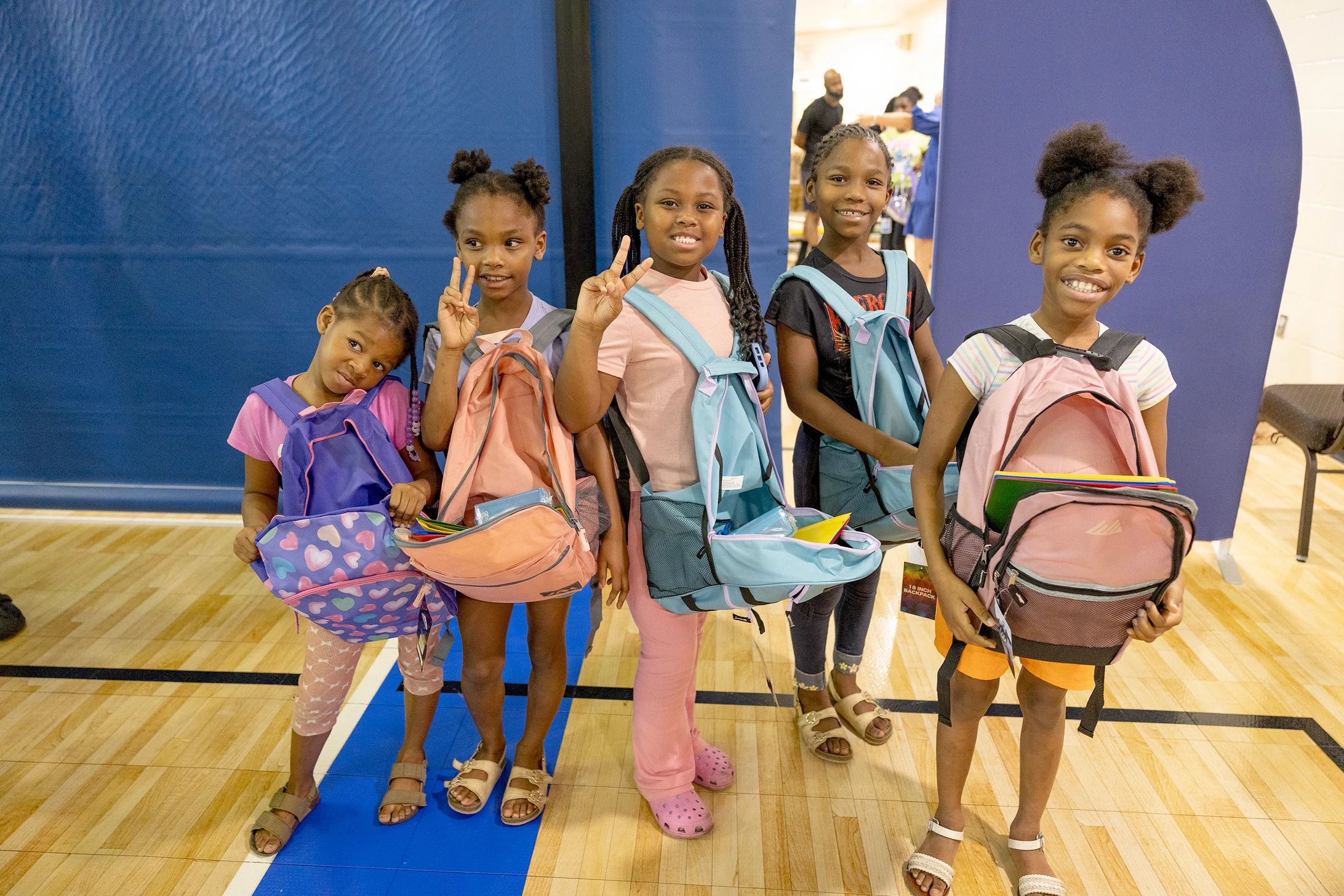 Five young Black girls stand in a gym, smiling and holding new backpacks filled with school supplies. Two of the girls are making peace signs with their hands.
