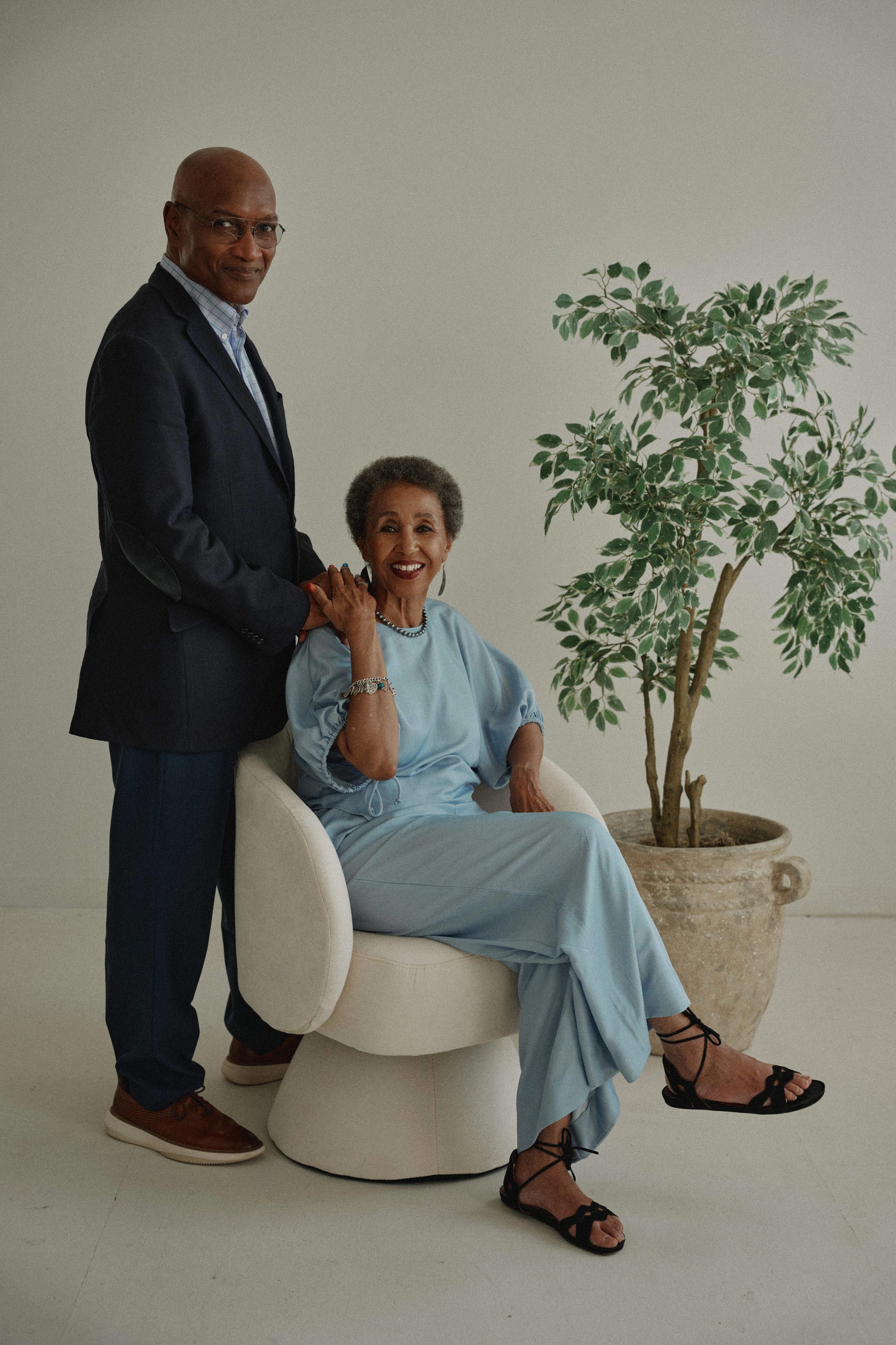 An elegantly dressed older Black couple poses for a portrait. The woman sits smiling in a chair, while the man stands behind her with his hand on her shoulder.