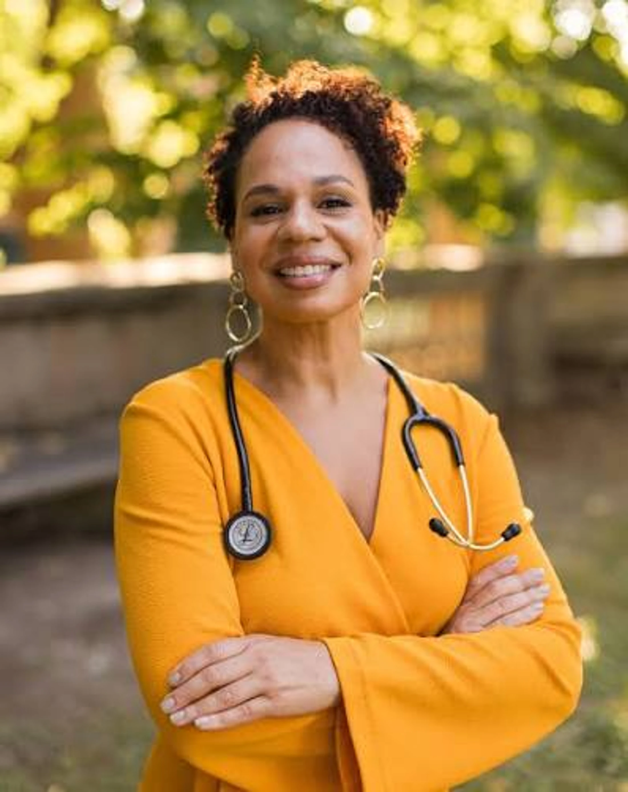 A smiling Black female doctor with a stethoscope around her neck stands outdoors with her arms crossed, wearing a bright yellow top.