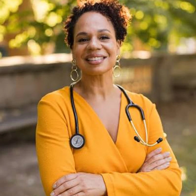 A smiling Black female doctor with a stethoscope around her neck stands outdoors with her arms crossed, wearing a bright yellow top.