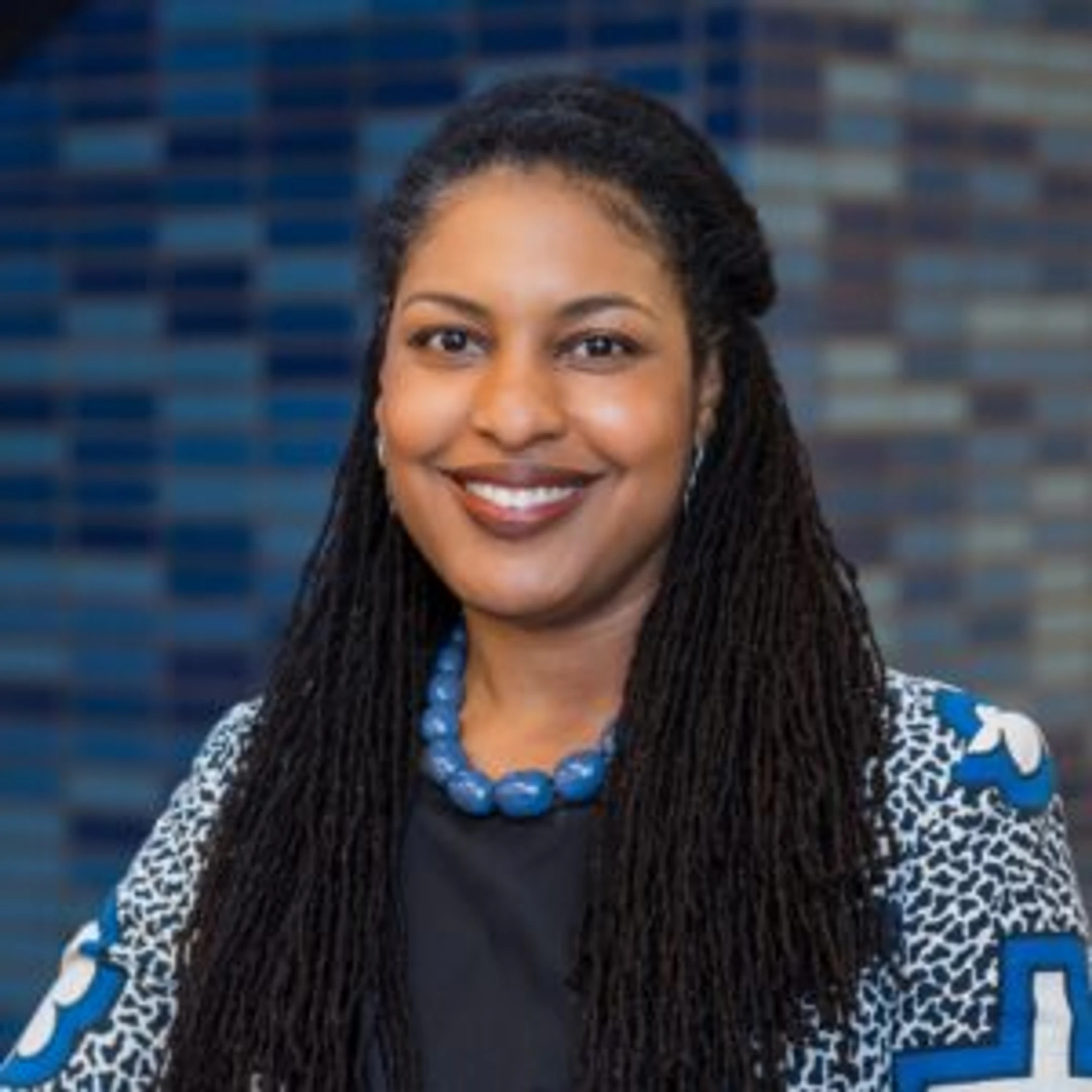 A professional headshot of a smiling Black woman with long locs, wearing a patterned jacket and a chunky blue necklace in front of a blue tiled wall.