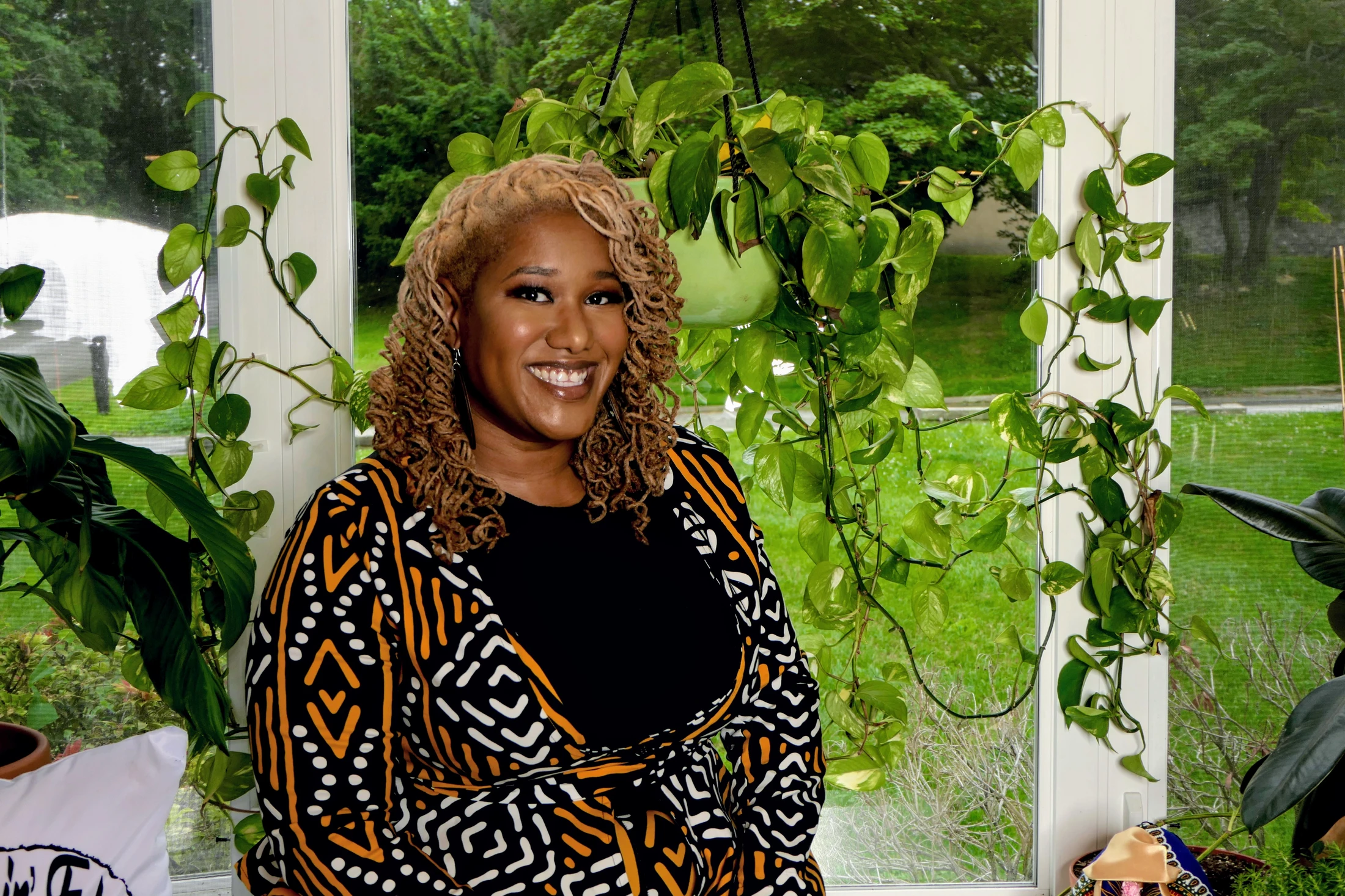 A Black woman with blonde curly hair smiles while wearing a patterned jacket. She stands in front of a large window, surrounded by vibrant green houseplants.