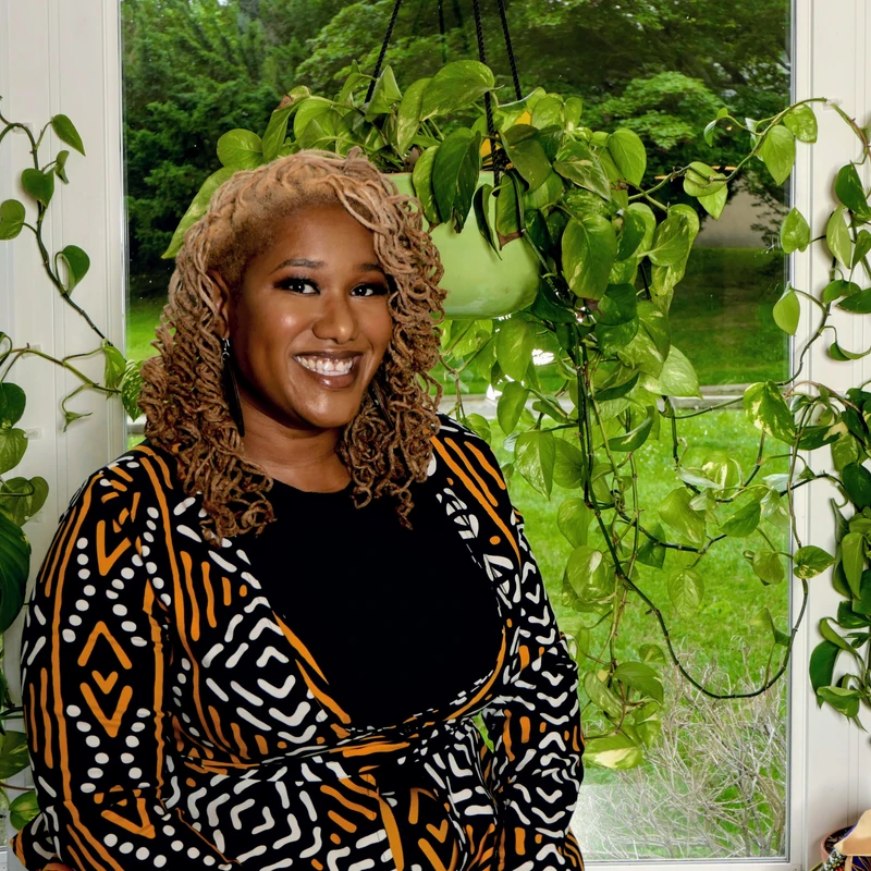 A Black woman with blonde curly hair smiles while wearing a patterned jacket. She stands in front of a large window, surrounded by vibrant green houseplants.