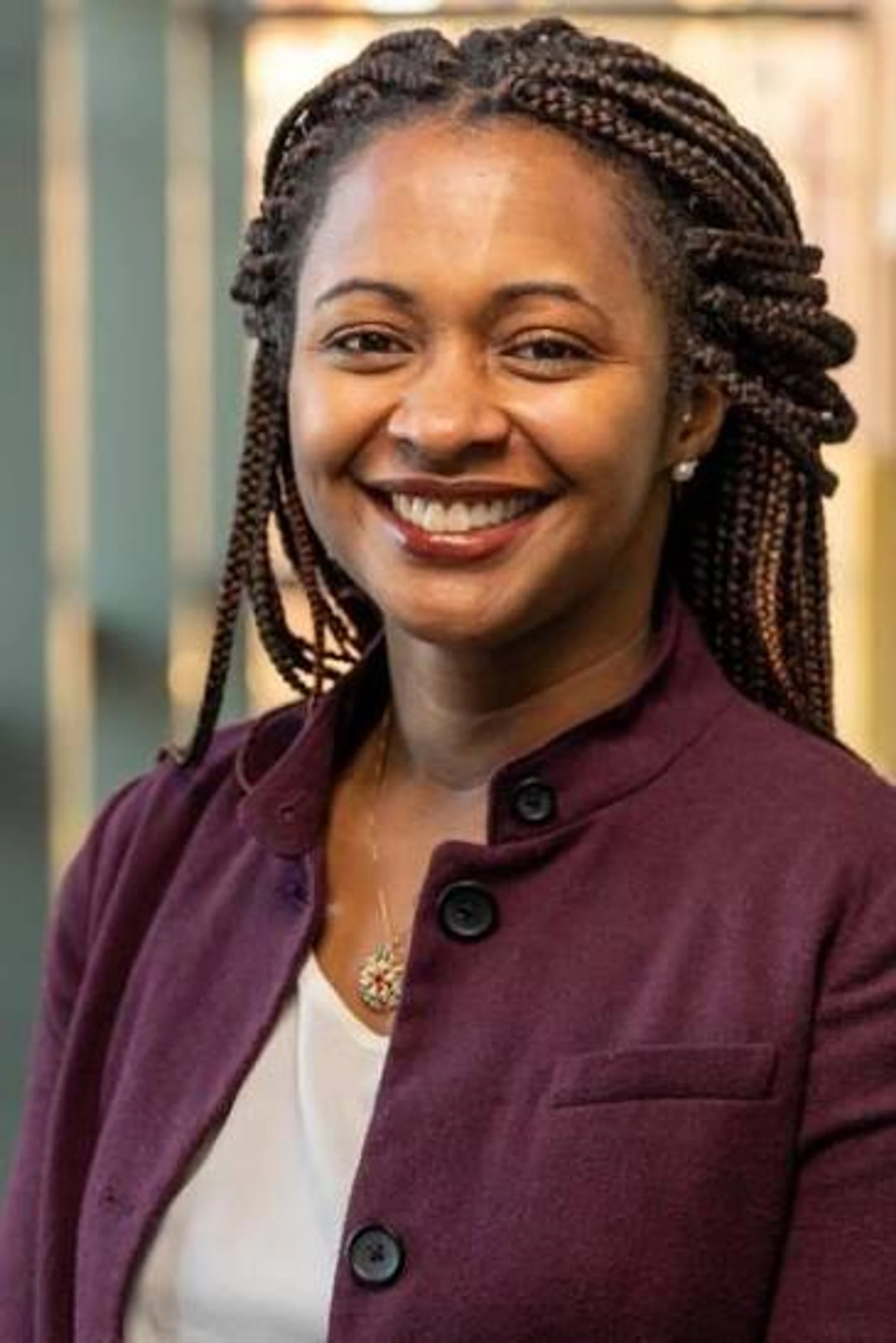 A headshot of a smiling Black woman with long braids. She is wearing a purple jacket over a white shirt and a delicate gold necklace.