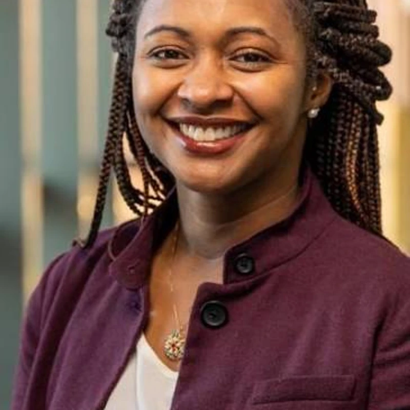 A headshot of a smiling Black woman with long braids. She is wearing a purple jacket over a white shirt and a delicate gold necklace.