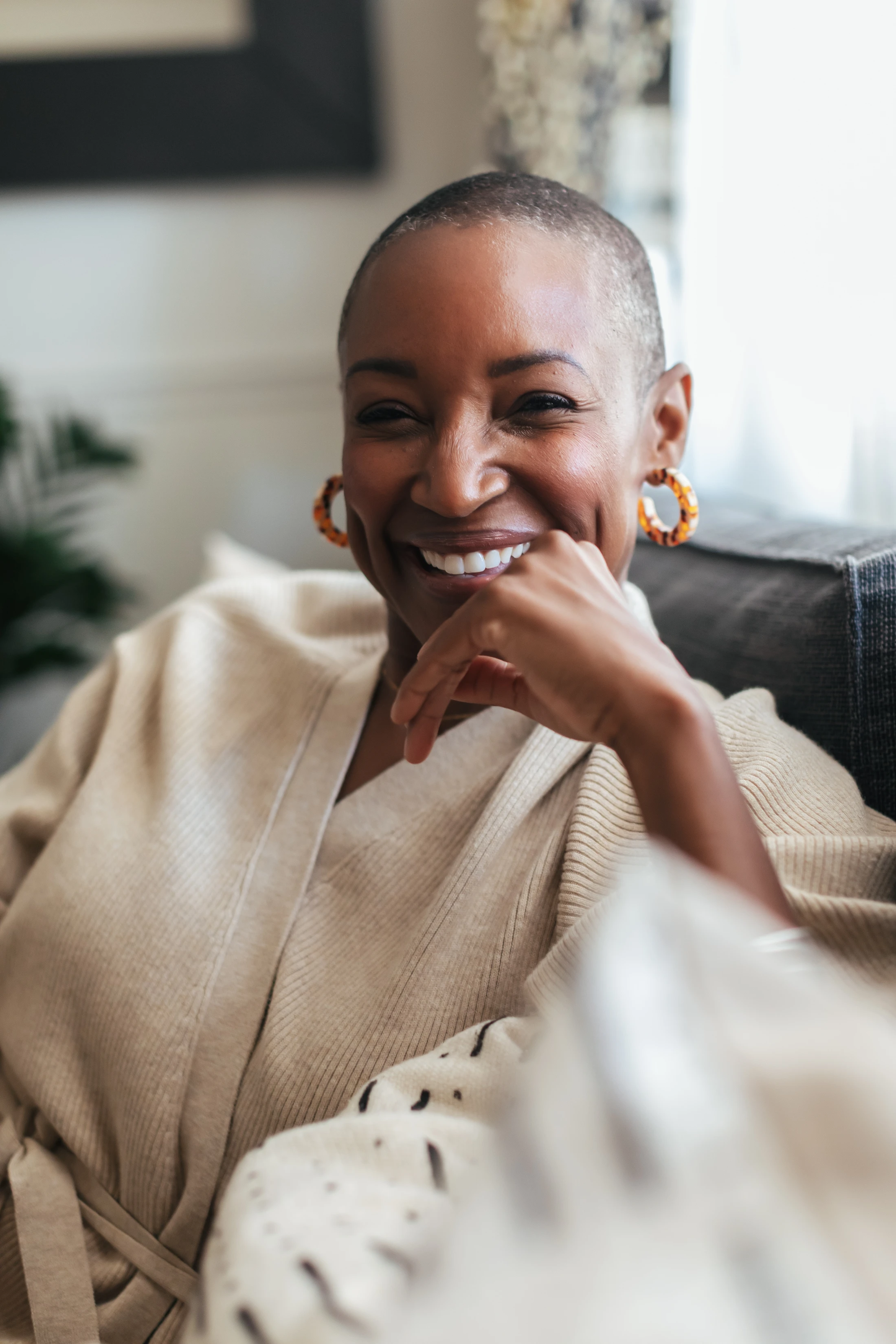 A close-up portrait of a smiling Black woman with a shaved head, wearing a cozy beige cardigan and hoop earrings, resting her chin on her hand.