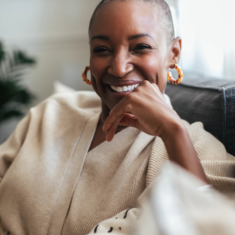 A close-up portrait of a smiling Black woman with a shaved head, wearing a cozy beige cardigan and hoop earrings, resting her chin on her hand.
