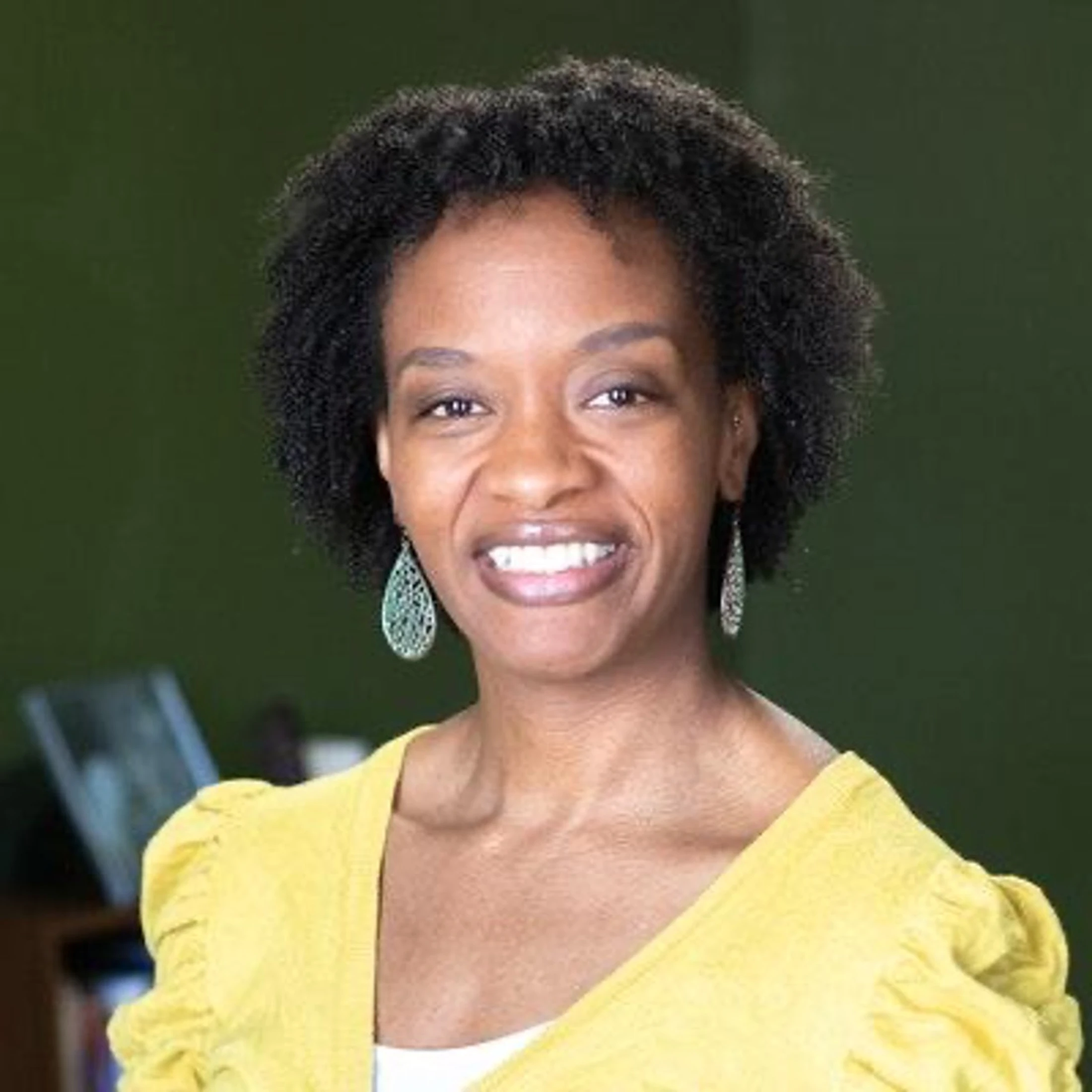 A professional headshot of a smiling Black woman with short curly hair. She wears a yellow cardigan, a white top, and large decorative earrings.