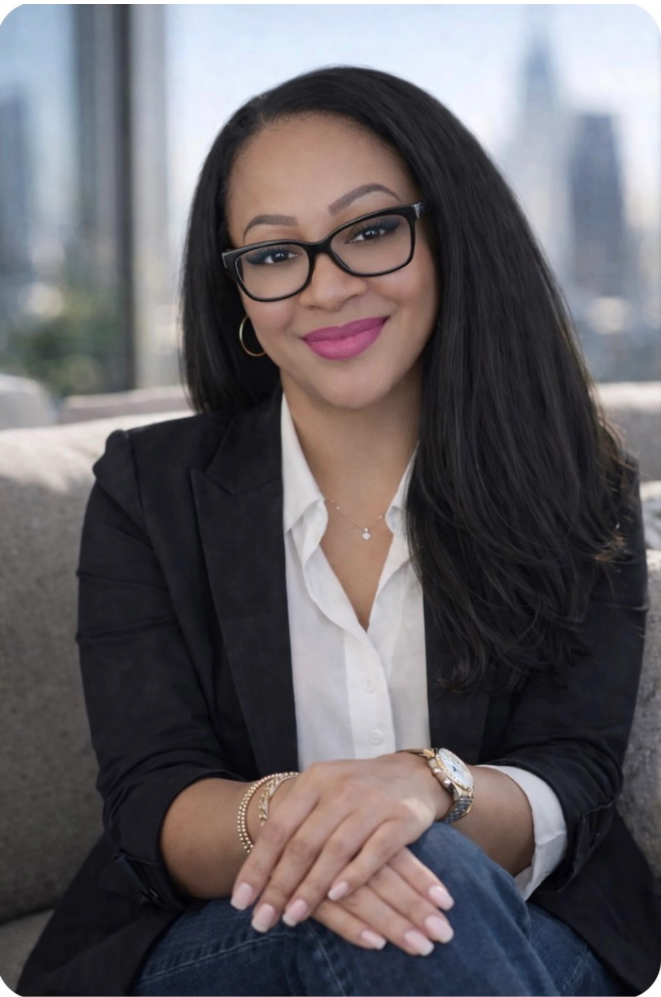A smiling woman with long dark hair and glasses sits on a couch. She wears a black blazer over a white shirt, with a blurred city skyline behind her.