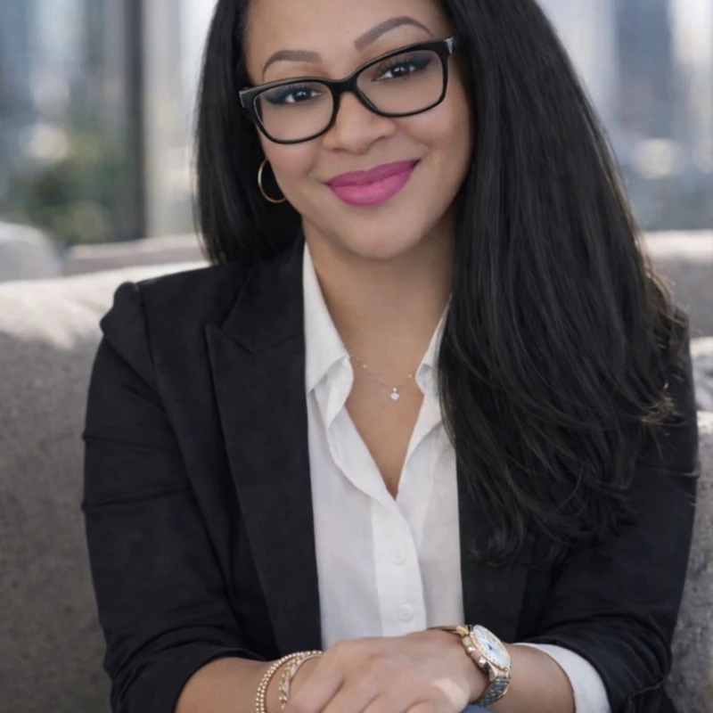 A smiling woman with long dark hair and glasses sits on a couch. She wears a black blazer over a white shirt, with a blurred city skyline behind her.