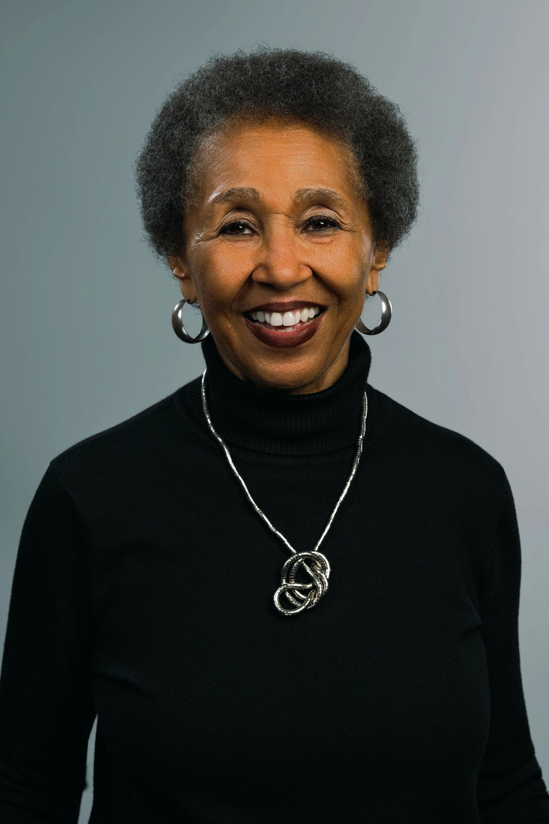 A headshot of a smiling older Black woman with short hair, wearing a black turtleneck, silver hoop earrings, and a silver necklace against a gray background.