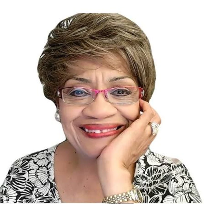 A smiling older woman with short brown hair and pink glasses poses for a headshot, resting her chin on her hand against a white background.