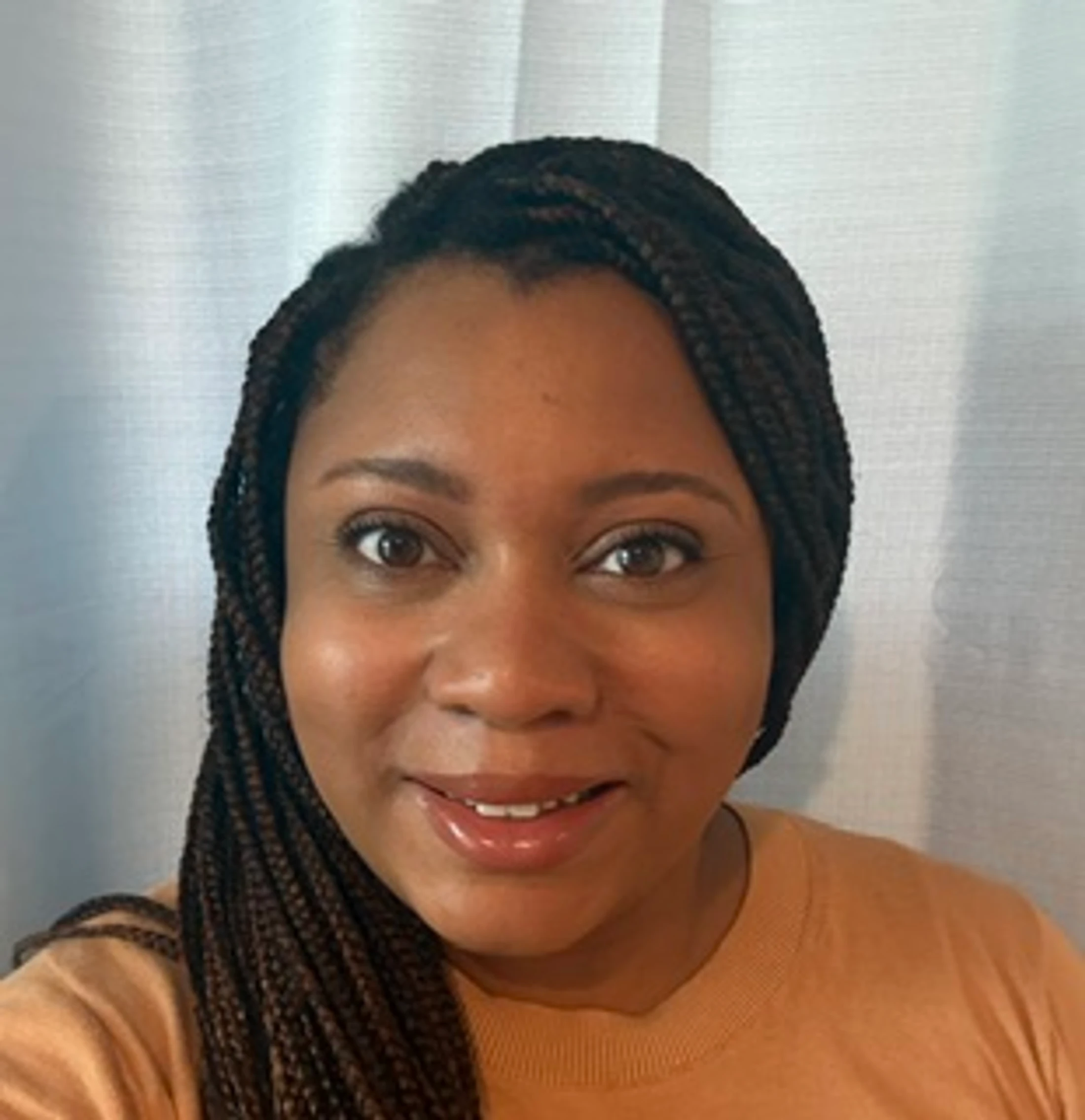 A head-and-shoulders portrait of a Black woman with long, dark braids. She is smiling and looking at the camera against a light curtain background.
