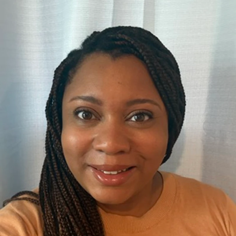 A head-and-shoulders portrait of a Black woman with long, dark braids. She is smiling and looking at the camera against a light curtain background.