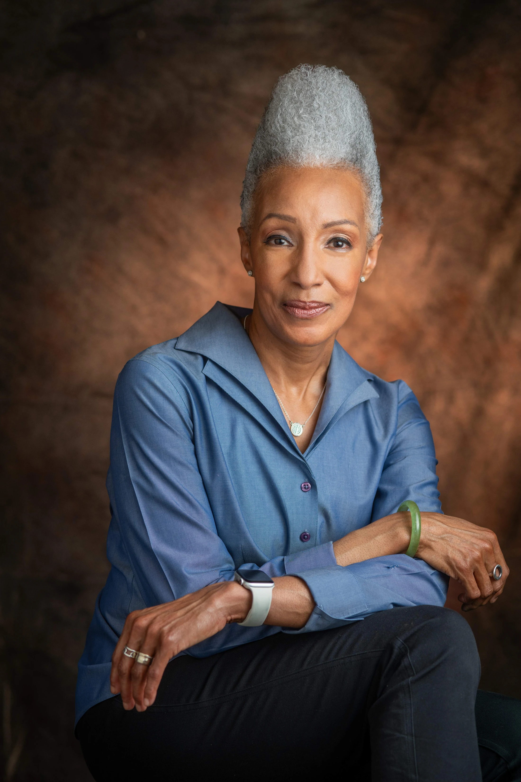 A professional studio portrait of an older Black woman with gray hair in an updo, wearing a blue shirt and smiling gently at the camera with her arms crossed.