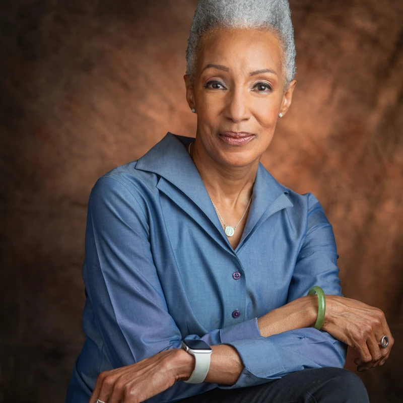 A professional studio portrait of an older Black woman with gray hair in an updo, wearing a blue shirt and smiling gently at the camera with her arms crossed.