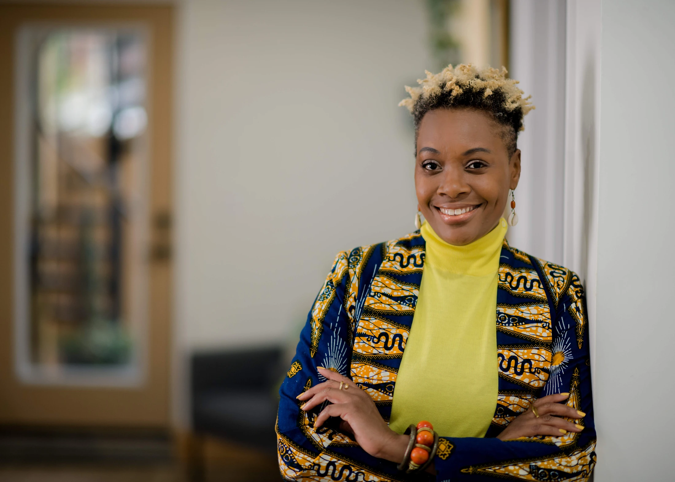 A smiling Black woman with short, blond-tipped hair, wearing a yellow turtleneck and a blue and yellow patterned jacket, stands with her arms crossed.