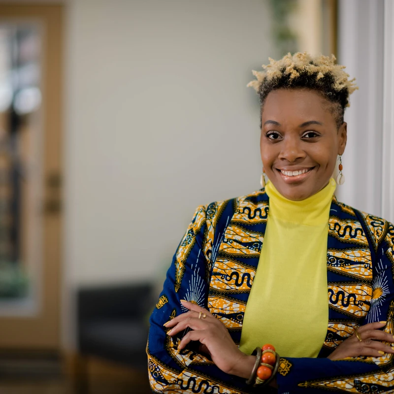 A smiling Black woman with short, blond-tipped hair, wearing a yellow turtleneck and a blue and yellow patterned jacket, stands with her arms crossed.