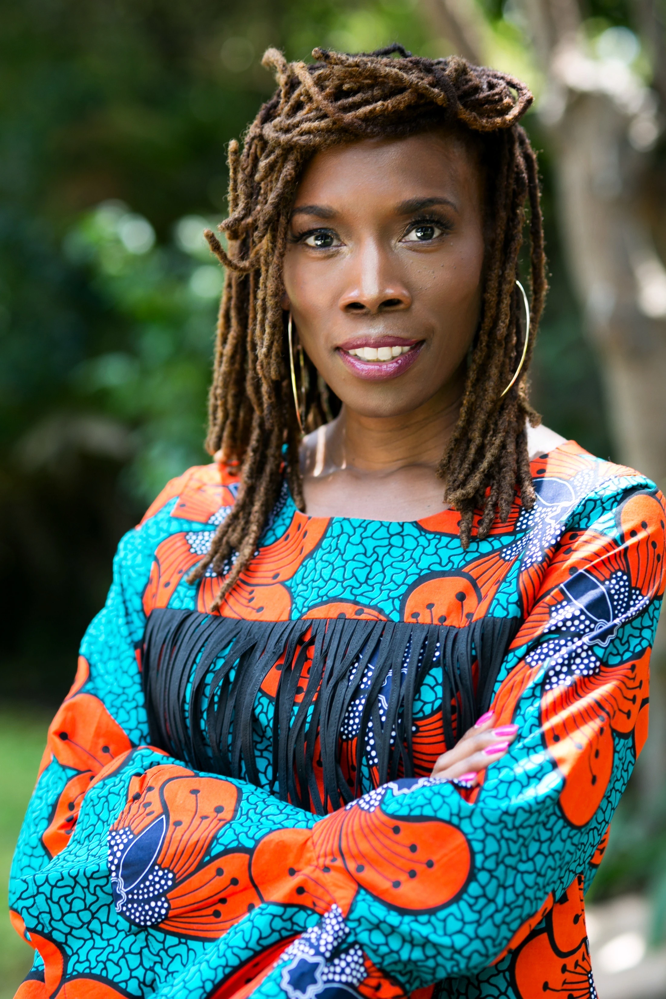 A portrait of a Black woman with brown locs, wearing a vibrant orange and turquoise African print top with black fringe. She stands outside with arms crossed.