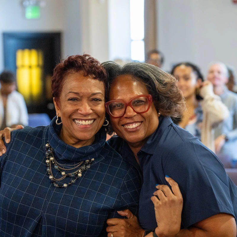 Two Black women smile warmly for the camera while embracing. They are indoors in an auditorium with rows of seats and other people in the blurred background.