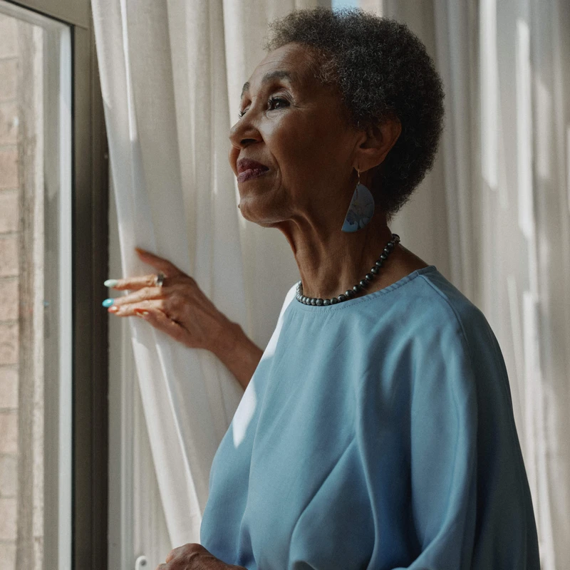 A poised elderly Black woman with short gray hair stands in profile, looking out a window and holding a white curtain, a gentle smile on her face.