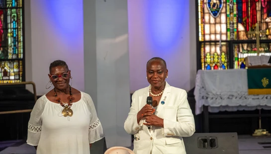 Two Black women in white outfits stand on a stage in a church. The woman on the right holds a microphone and speaks, while the other stands beside her.
