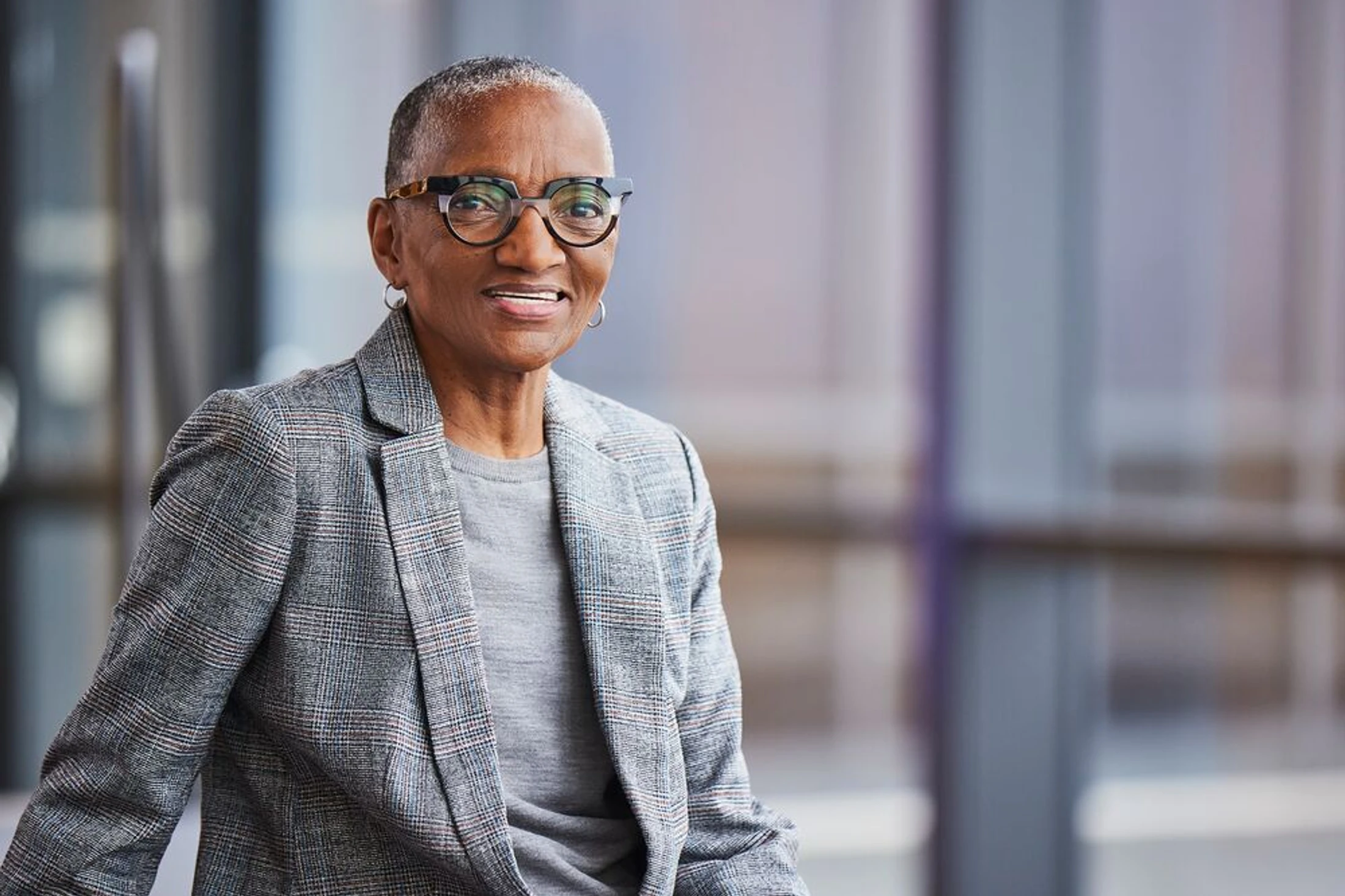 A smiling senior Black woman with short hair and stylish glasses, wearing a gray plaid blazer and looking directly at the camera in a modern office.