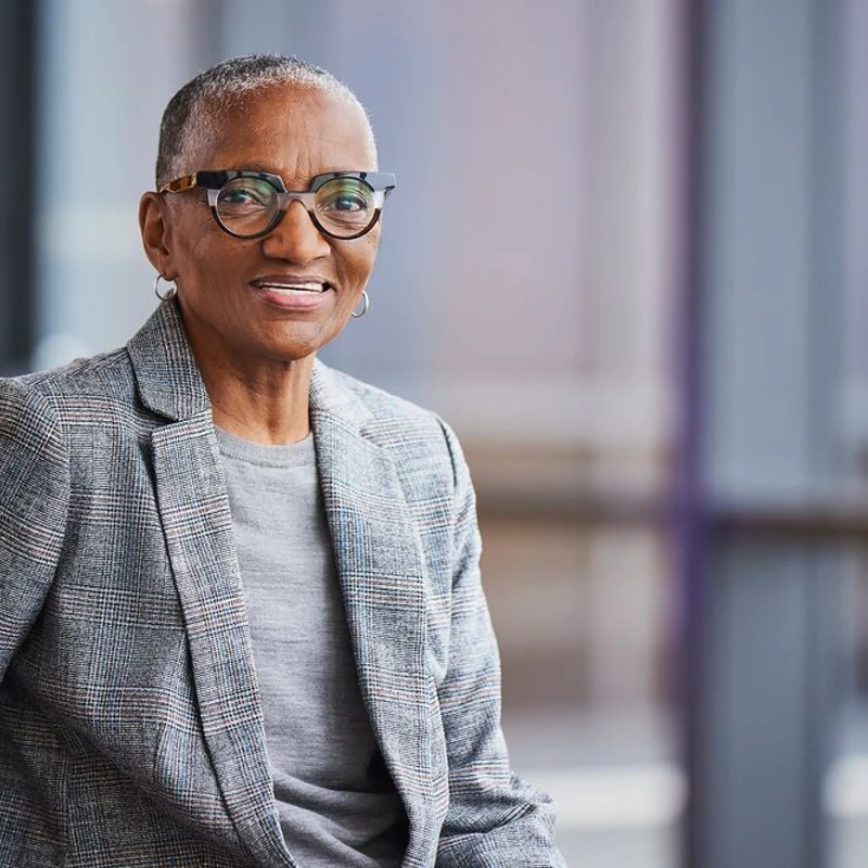 A smiling senior Black woman with short hair and stylish glasses, wearing a gray plaid blazer and looking directly at the camera in a modern office.