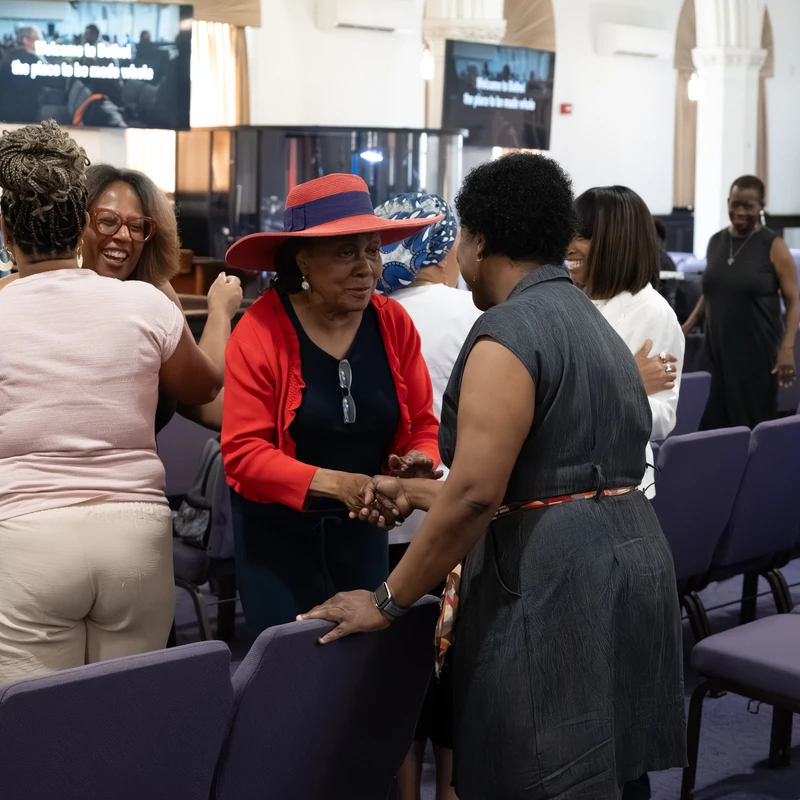 A group of Black women warmly greet each other among rows of chairs in a church. A woman in a red hat shakes hands with another while others smile.
