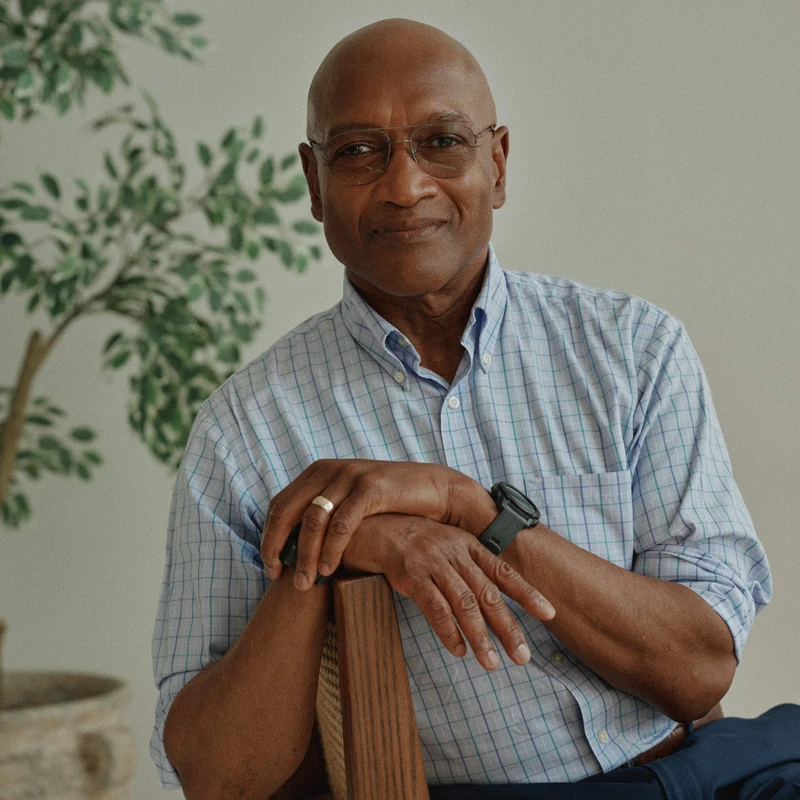 A bald, older Black man with glasses smiles warmly at the camera. He wears a plaid shirt and sits backwards on a wooden chair with his arms resting on top.