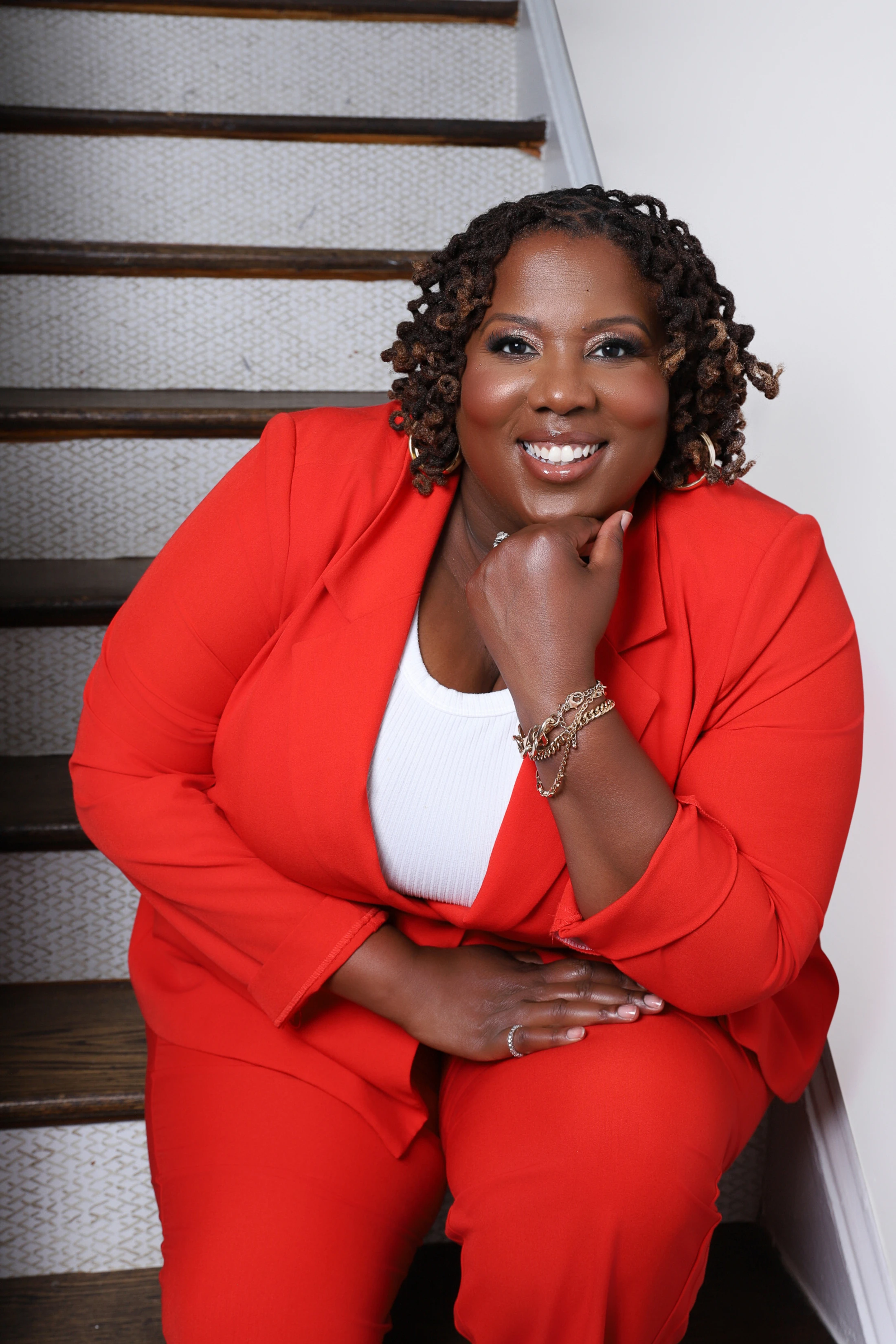 A smiling Black woman with locs wears a bright red pantsuit while sitting on a staircase. She rests her chin on her hand and looks at the camera.