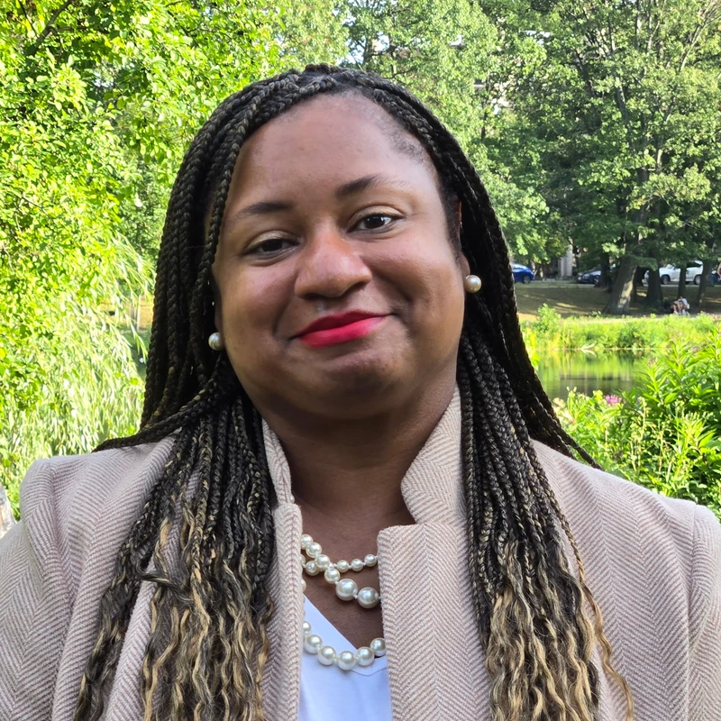 A headshot of a smiling Black woman with long, ombre braids. She wears a beige blazer, a pearl necklace, and red lipstick while standing in a park.