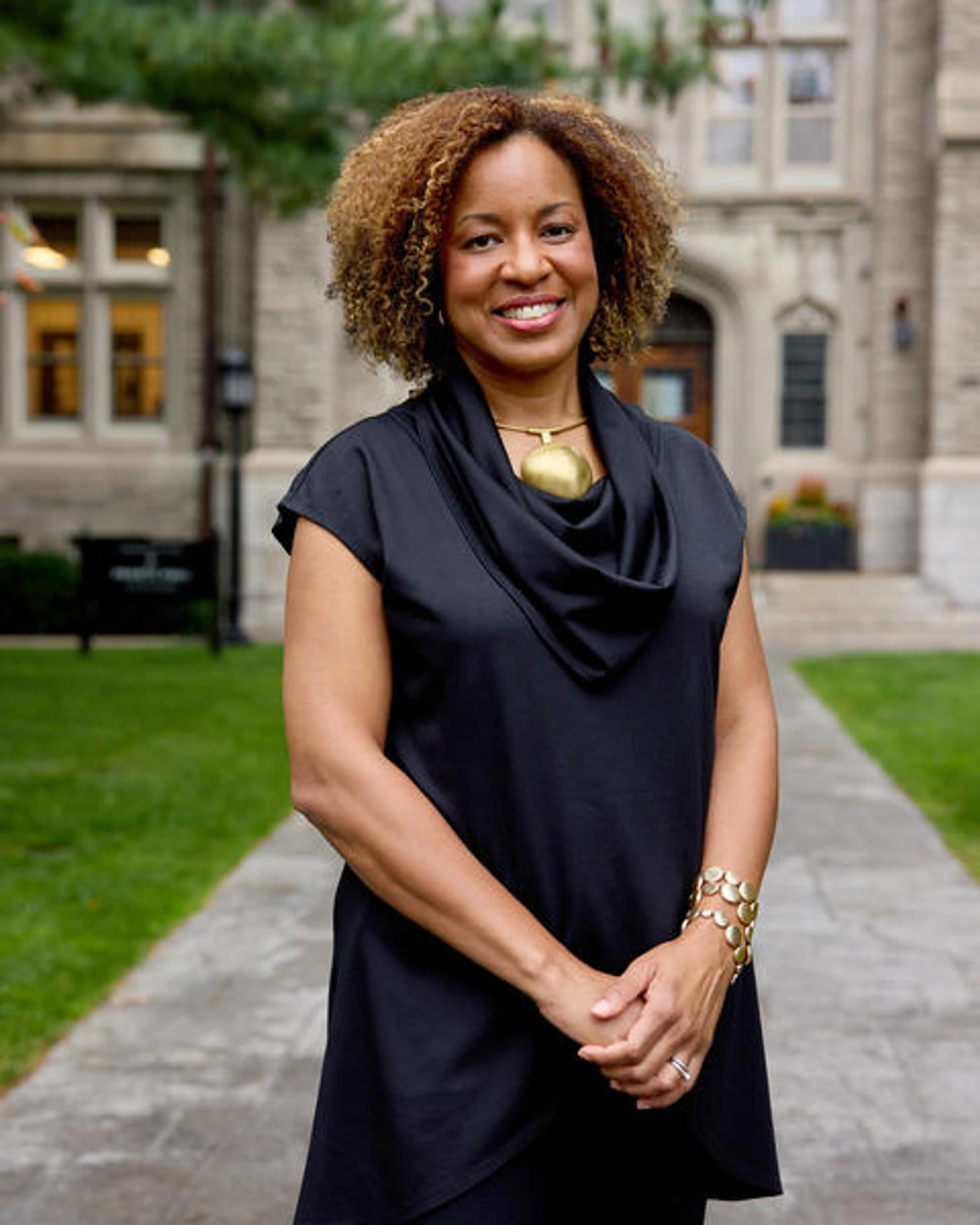 A smiling woman with curly, highlighted hair stands on a paved path outdoors. She wears a black top and gold jewelry, with a large stone building behind her.
