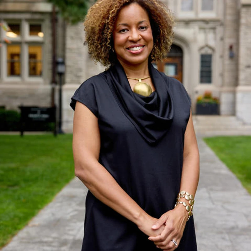 A smiling woman with curly, highlighted hair stands on a paved path outdoors. She wears a black top and gold jewelry, with a large stone building behind her.