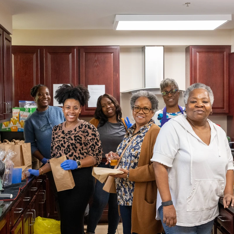 A group of six Black women of various ages smiling for the camera while standing in a kitchen, preparing food and drinks for a community event.