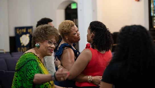 Several Black women gather indoors. Two women in the center embrace and talk intimately, while another woman in a bright green top smiles in the foreground.
