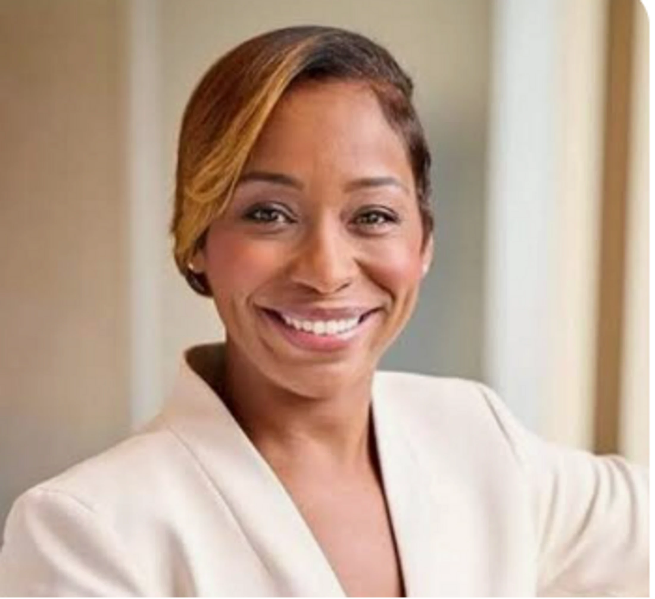 A headshot of a smiling Black woman with short, highlighted hair, wearing a light-colored professional blazer against a blurred indoor background.