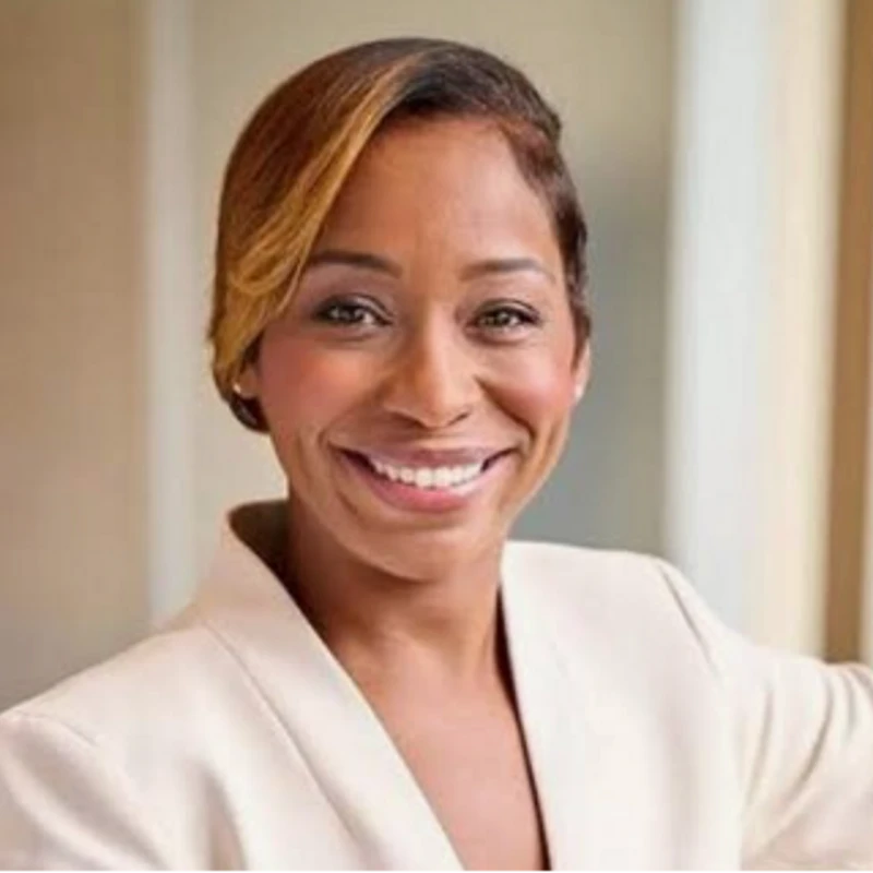 A headshot of a smiling Black woman with short, highlighted hair, wearing a light-colored professional blazer against a blurred indoor background.