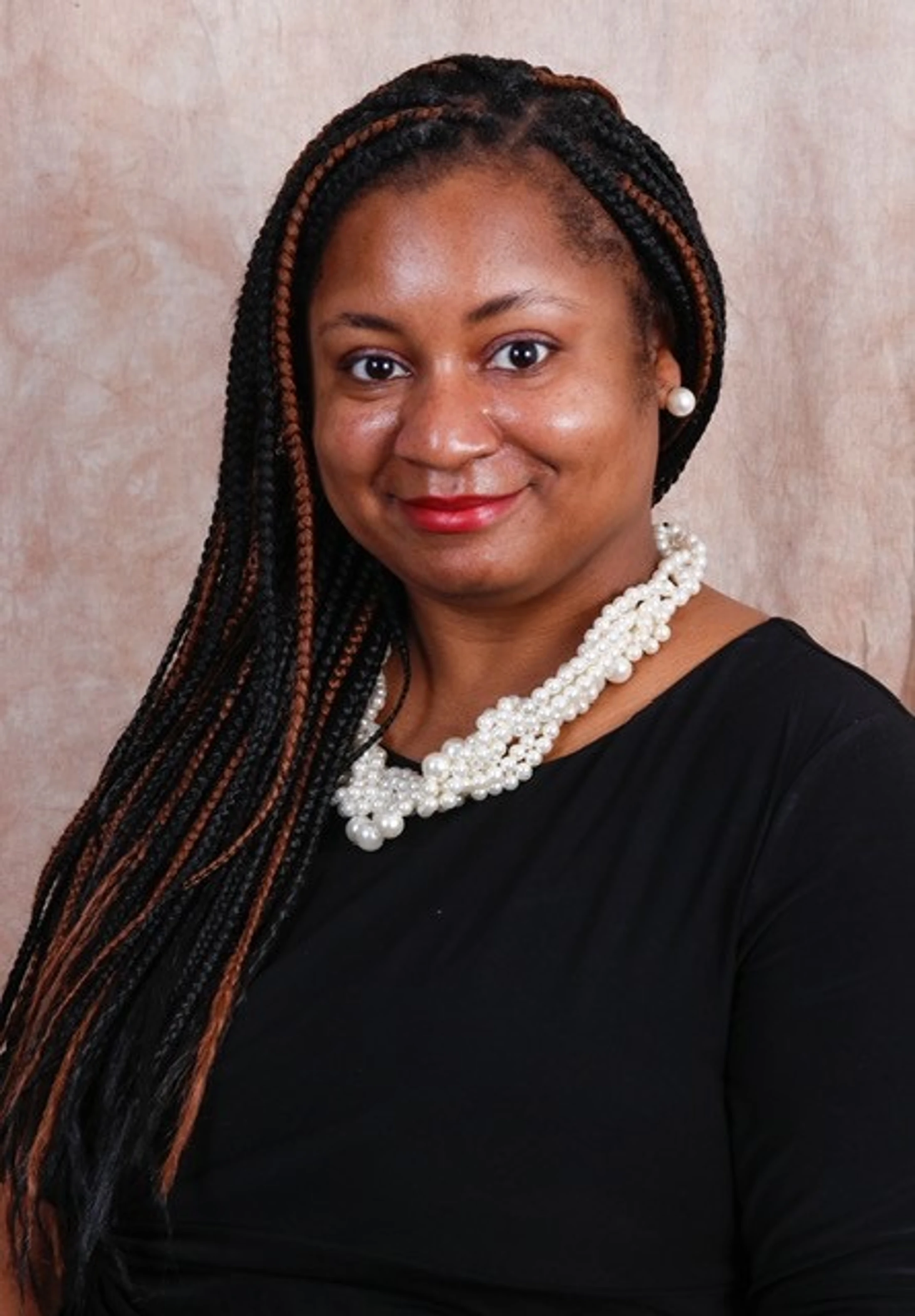 A smiling Black woman with long black and brown box braids, wearing a black shirt, a multi-strand pearl necklace, and pearl earrings against a brown backdrop.