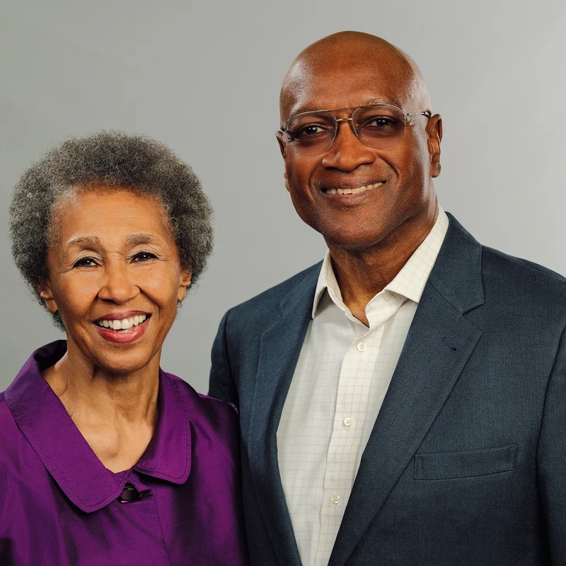 A warmly smiling older Black woman in a purple jacket stands next to a smiling bald Black man in a suit and glasses against a gray background.