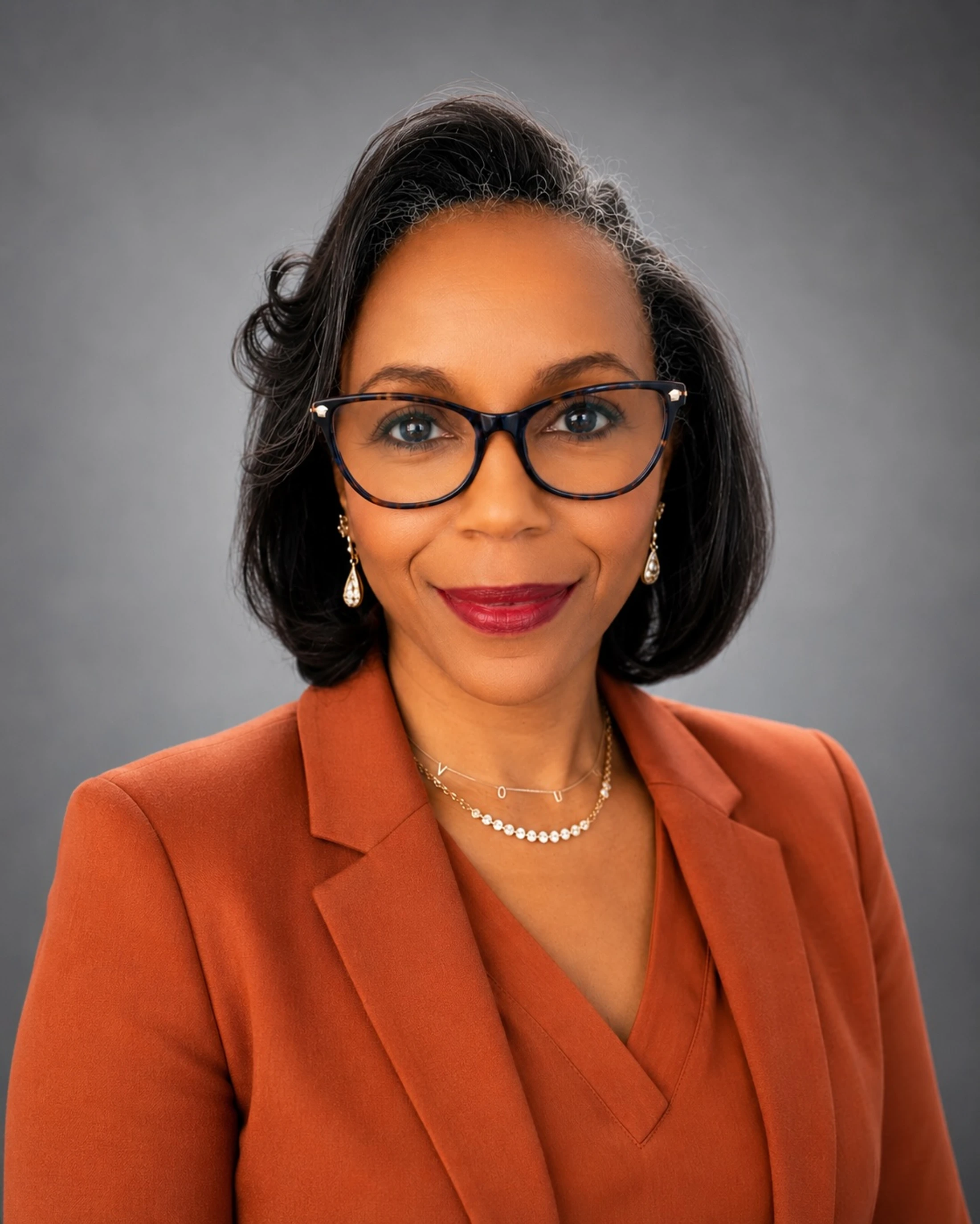 A professional headshot of a smiling Black woman with short dark hair and glasses, wearing a rust-colored blazer against a plain gray background.