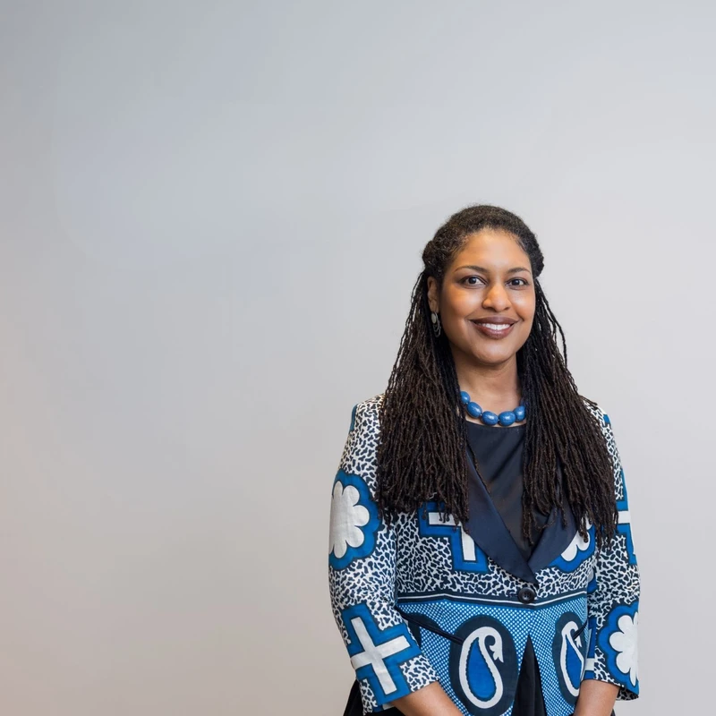 A smiling woman with long dreadlocks stands against a plain background. She is wearing a distinctive blue and white patterned blazer and a blue beaded necklace.
