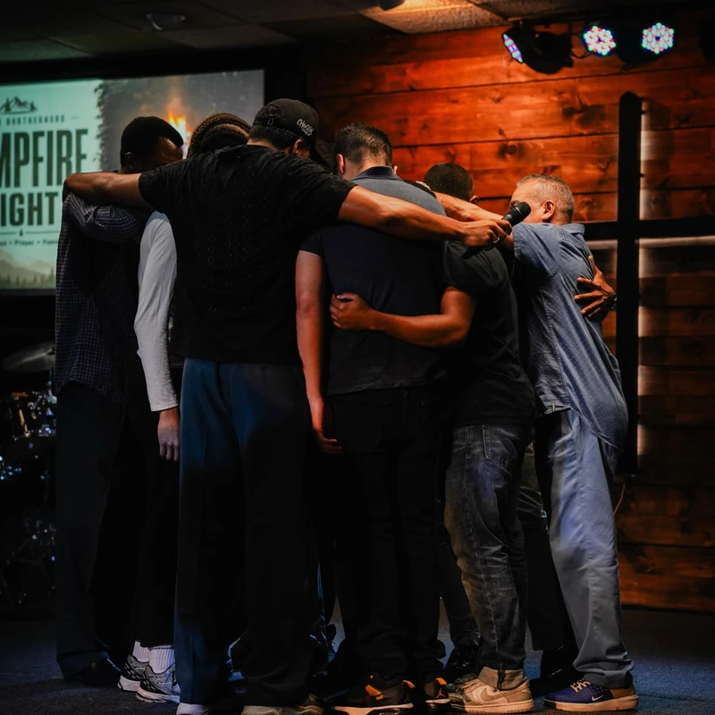 A diverse group of men stands in a tight circle on a stage, arms around each other in prayer or fellowship. A screen in the background reads 'Campfire Night'.